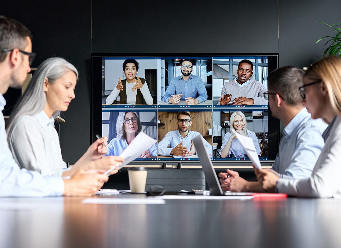 Insurance Solutions - Group of Business Professionals Sit Together at an Office With Other Members on the Screen Participating Virtually